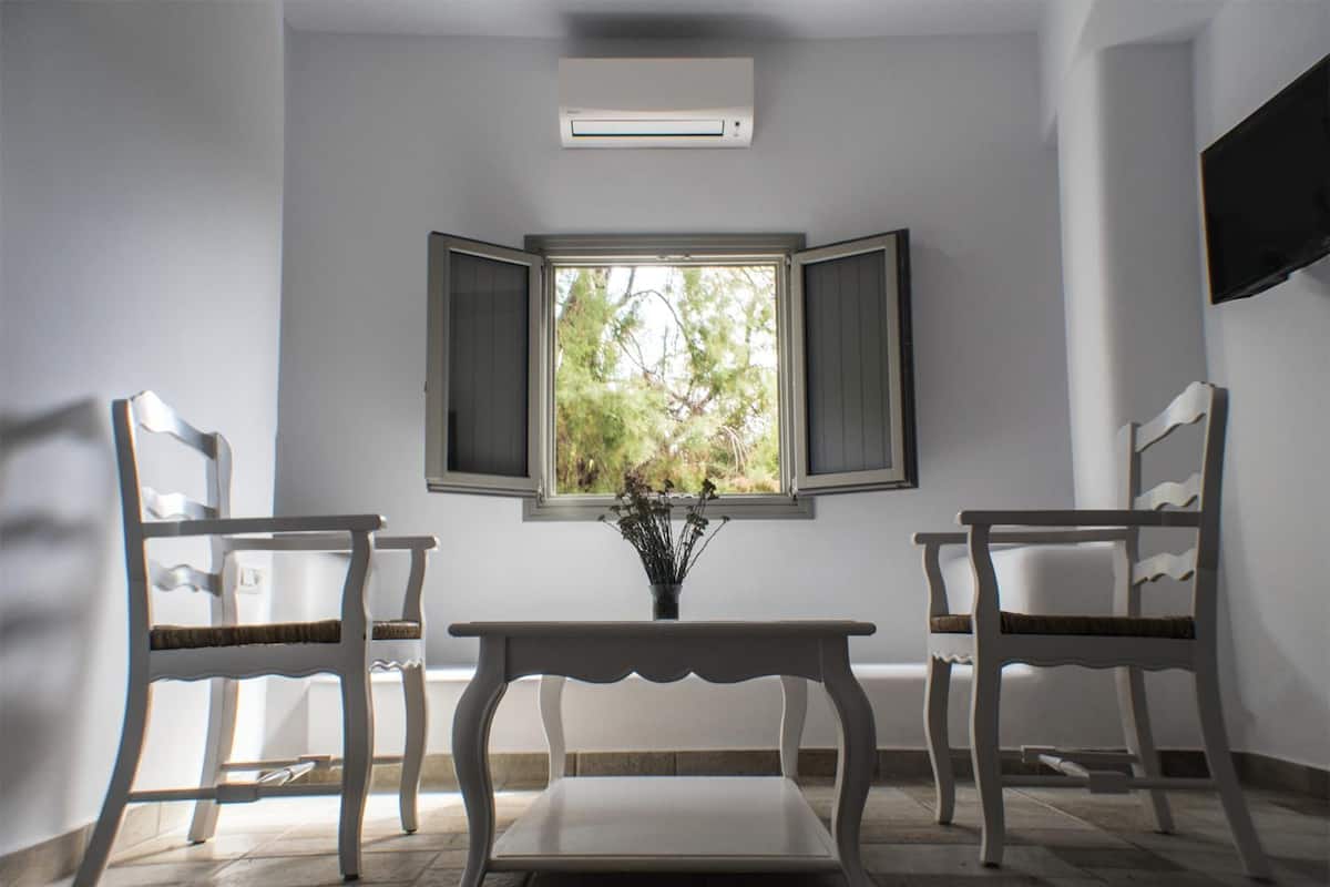 Quarto com duas cadeiras brancas, uma mesa pequena e uma janela com as persianas abertas.