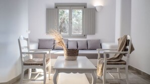 Une pièce avec un canapé blanc, deux fauteuils blancs, une petite table blanche et une fenêtre avec des volets.