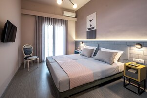 Una habitación de hotel moderna con una cama grande, una mesita de noche, una silla, un televisor y un ventanal con cortinas.
