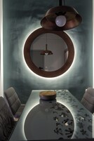 Un comedor moderno con una mesa de vidrio, sillas acolchadas, un espejo redondo e iluminación colgante.
