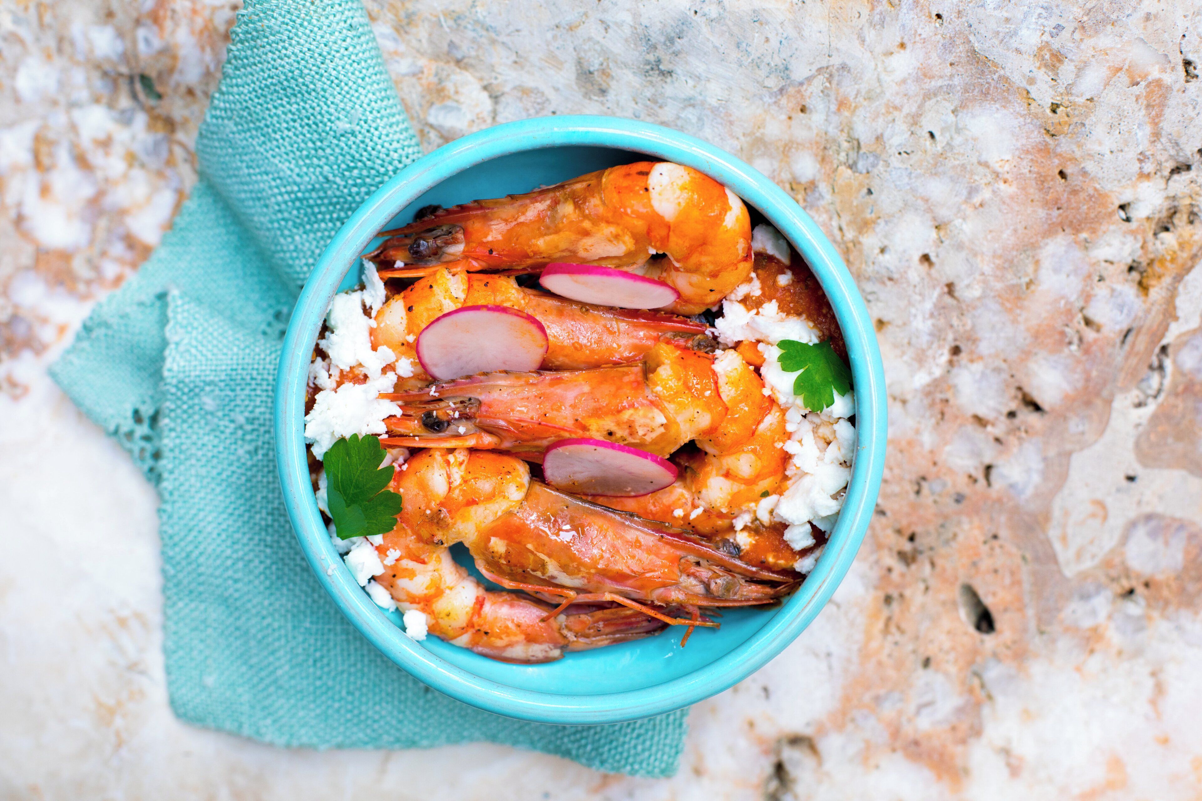 A bowl of cooked shrimp with radishes and herbs on a textured surface.