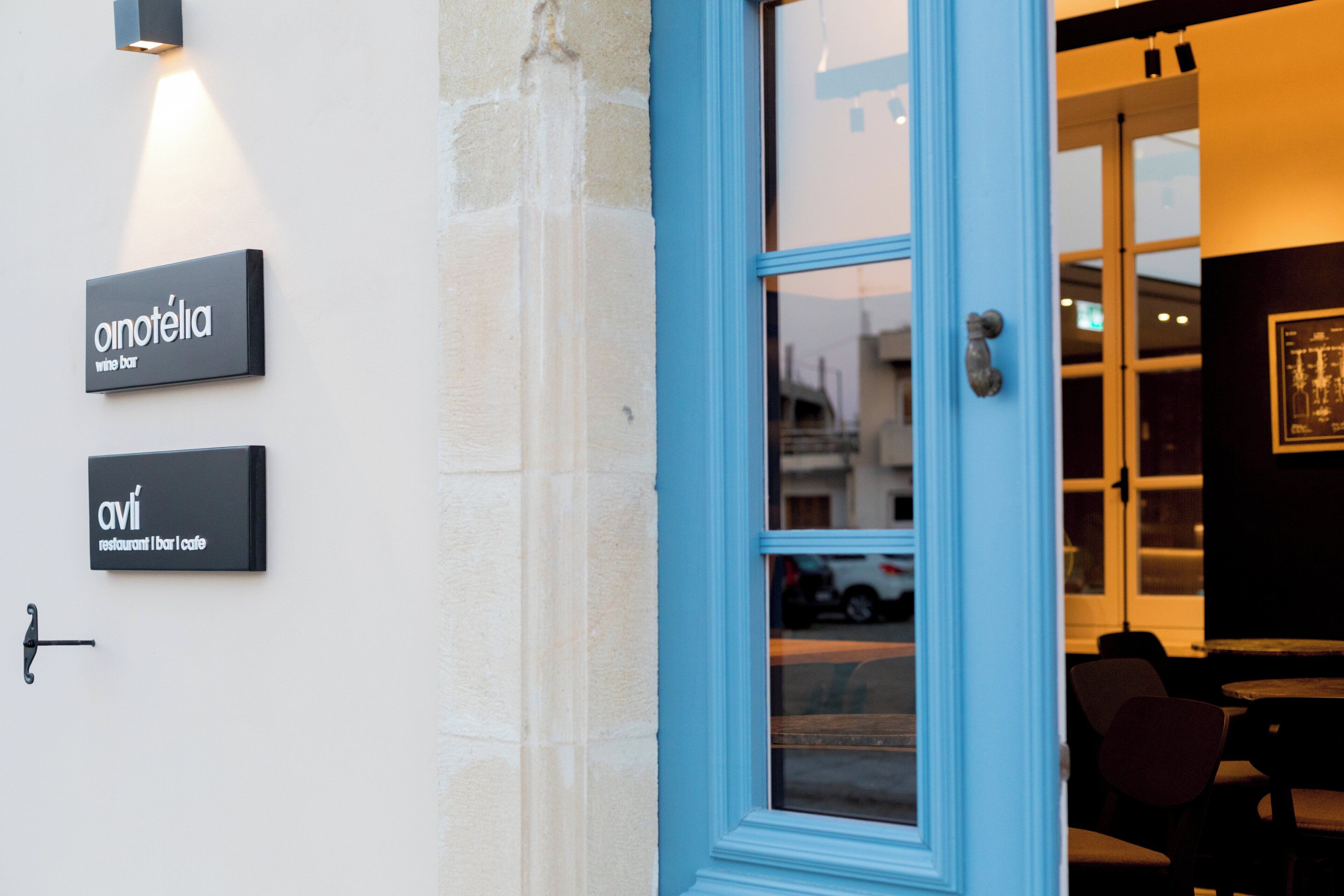 A building entrance with a blue door and two signs indicating 'onotélia' and 'avi' as a restaurant, bar, and cafe.