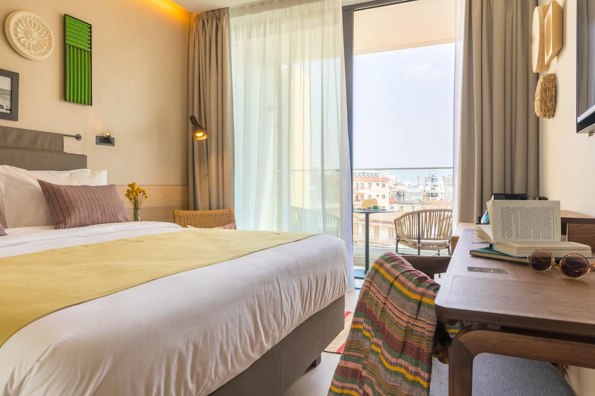 A hotel room with a bed, a desk with a chair, a window with a view of buildings, and a balcony.