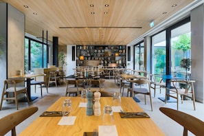 A modern dining area with wooden tables and chairs, a central display of decorative items, and large windows offering views of the outdoors. - Hotel Indigo Larnaca by IHG (Larnaca)