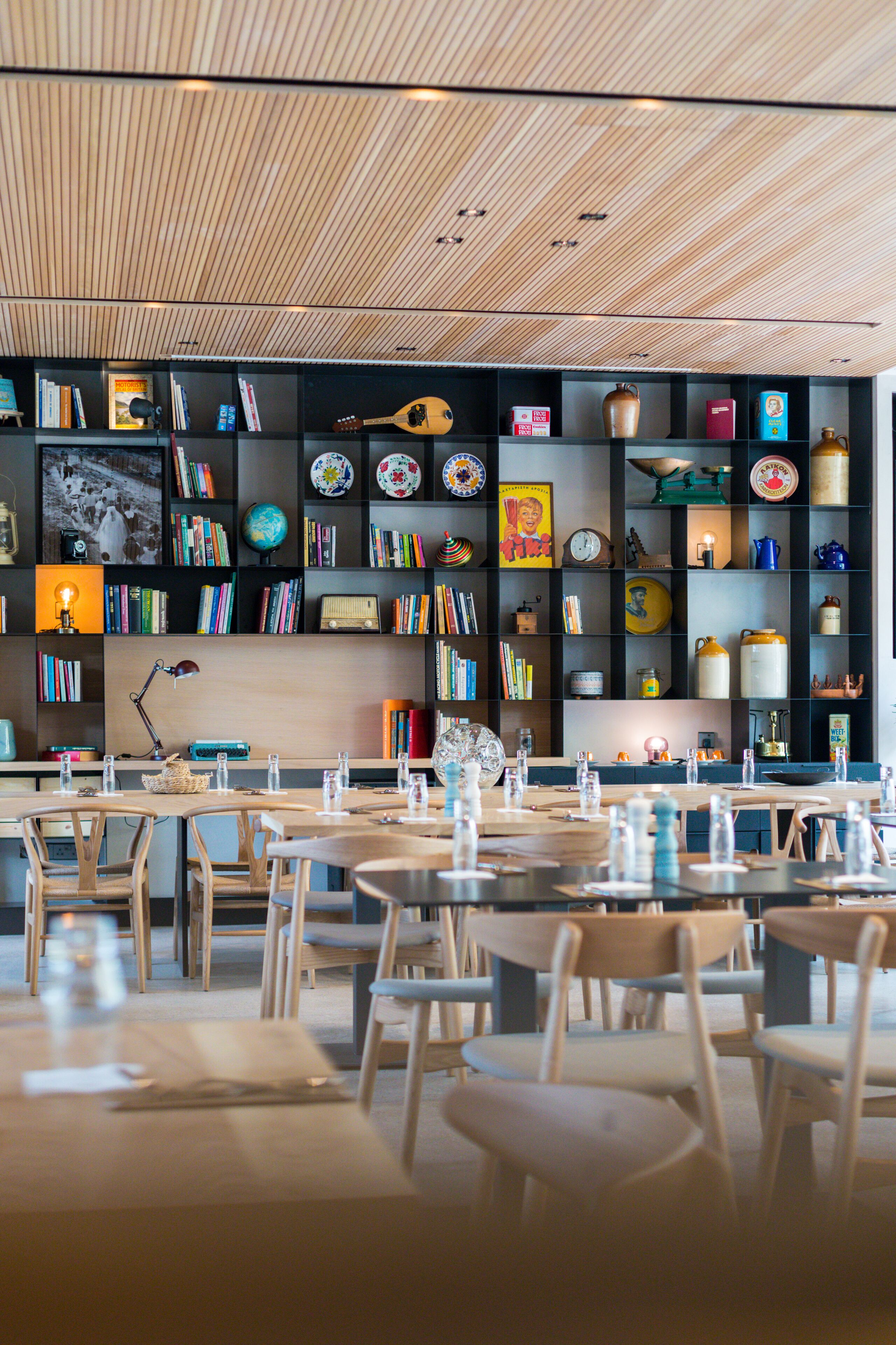 A modern dining area with wooden tables and chairs, a bookshelf filled with books and decorative items, and a wooden ceiling.