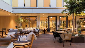 An outdoor seating area with wooden furniture and tables, surrounded by plants and trees.