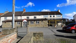 A two-story stone building with a traditional design, featuring a small entrance with a red door, a covered outdoor seating area, and a parked red car. - The Nags Head (Thirsk)