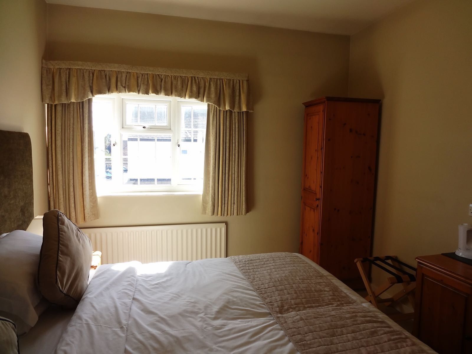 Une chambre à coucher avec un lit, un radiateur, une fenêtre avec des rideaux, une armoire en bois et une chaise.