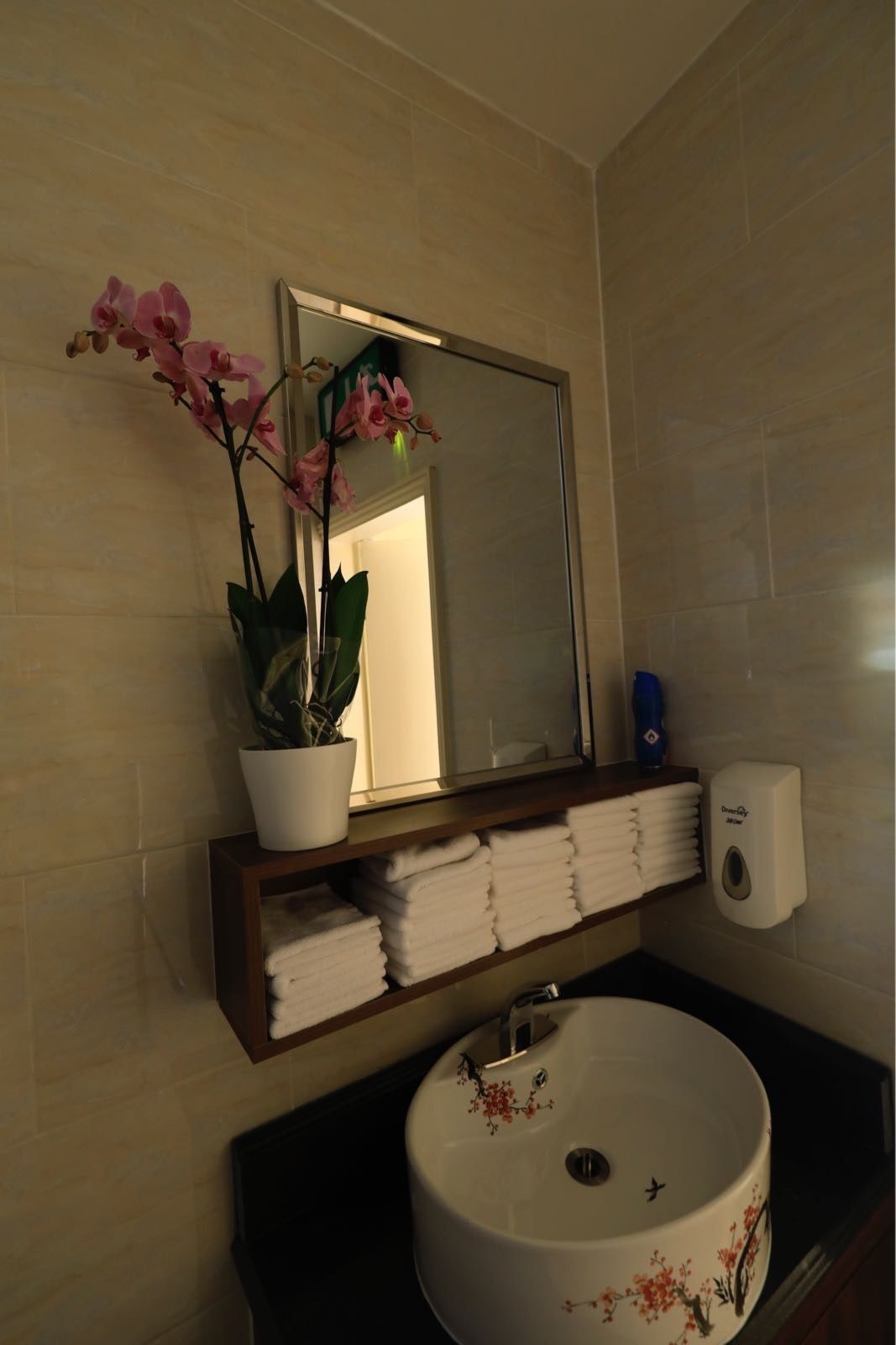 A bathroom with a white sink, a mirror, a potted orchid, and neatly folded towels on a shelf.