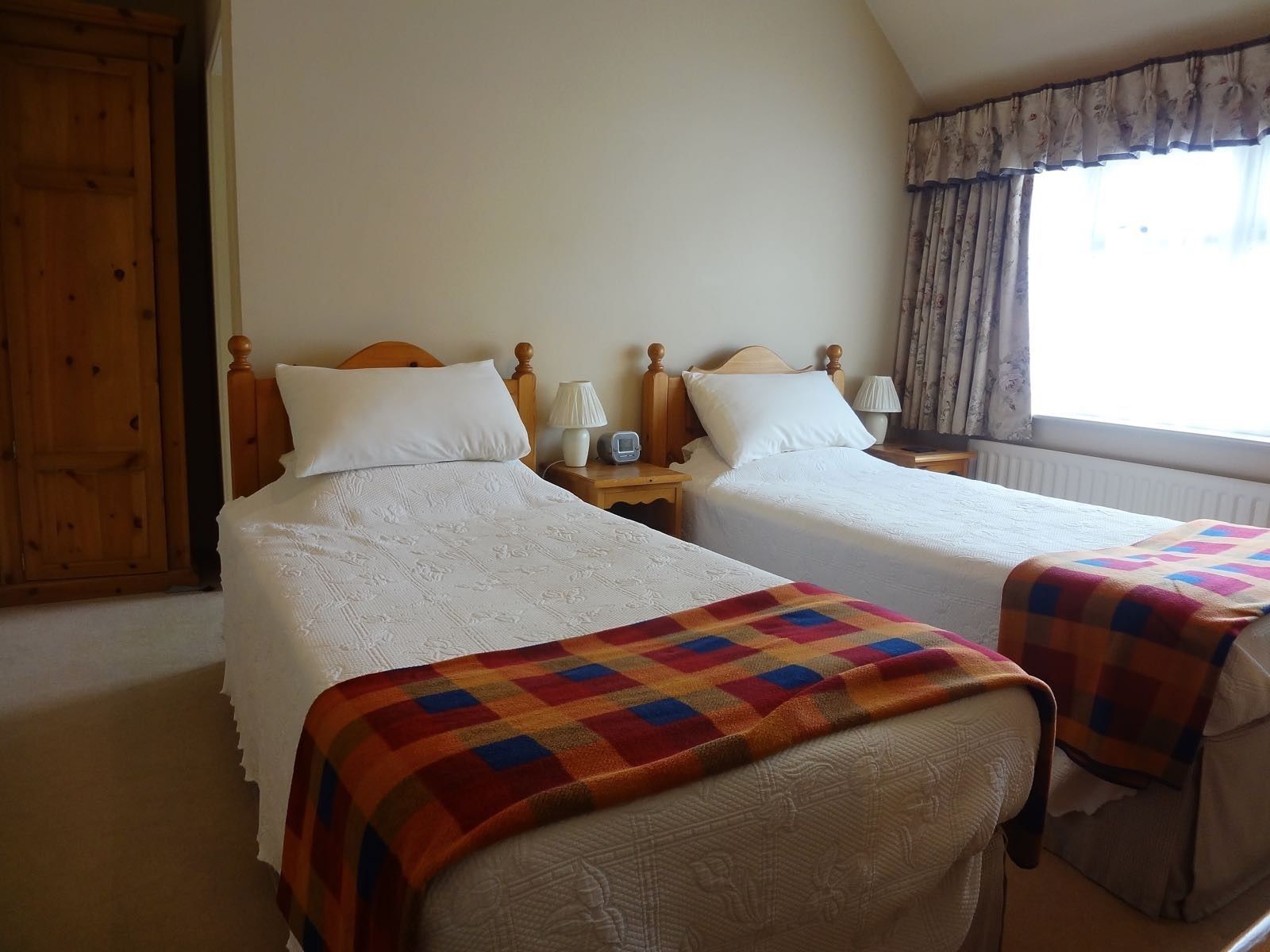 A hotel room with two beds, each with white linens and a colorful blanket on one bed. There is a wooden wardrobe, a nightstand with a lamp, and a window with curtains.