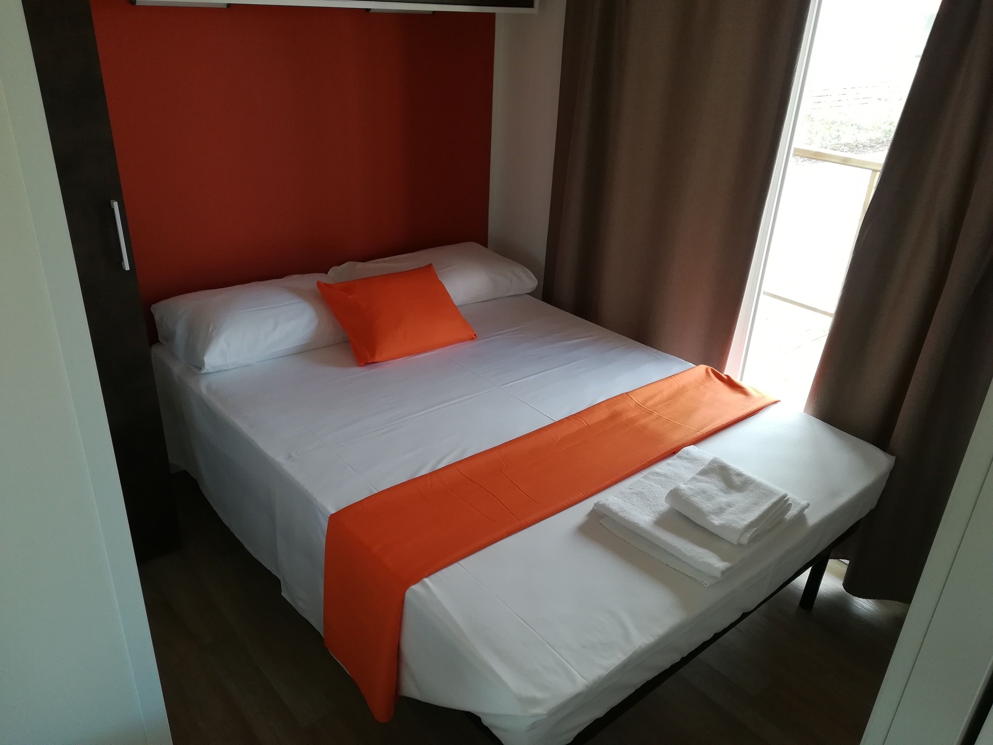 A neatly made bed with white linens and an orange throw blanket, a red accent wall, and a window with curtains.