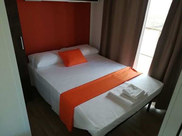 A neatly made bed with white linens and an orange throw blanket, a red accent wall, and a window with curtains. - Camping Verona Village (Verona)