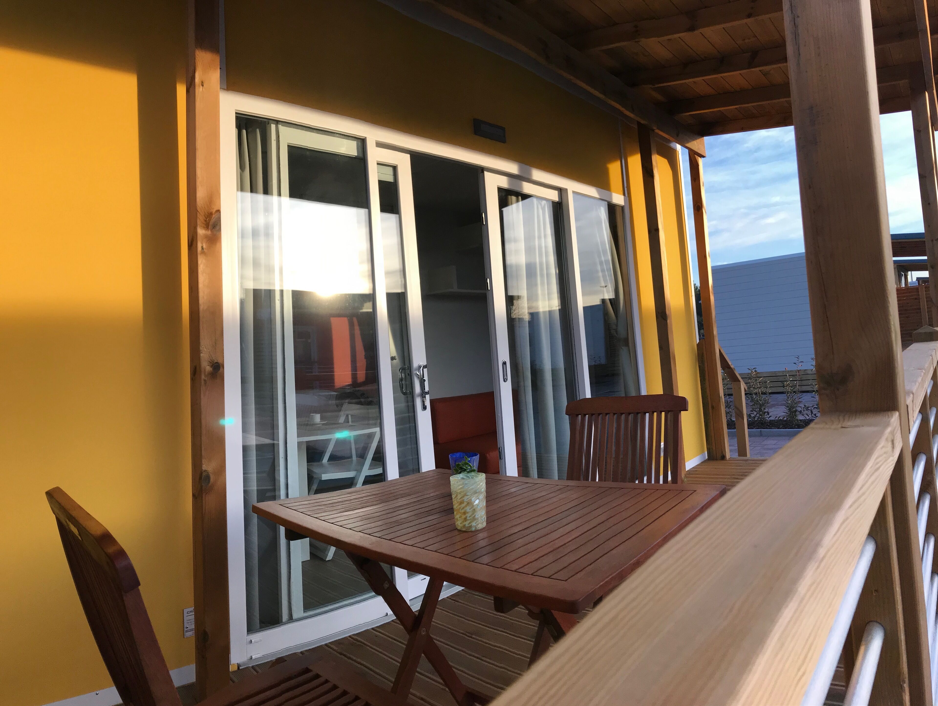 A wooden deck with a table and chairs, a sliding glass door leading to an interior space, and a view of the sea.