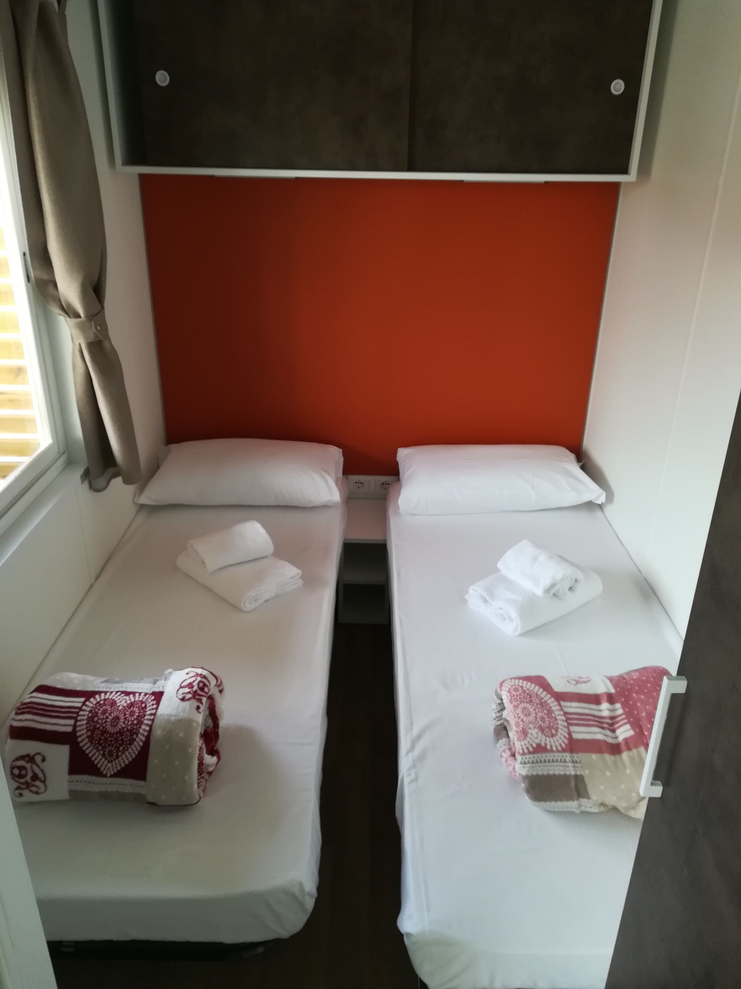 Una habitación pequeña y compacta con dos camas individuales, cada una con colchón y almohadas blancos. Hay toallas sobre las camas y una ventana con cortinas al lado izquierdo.