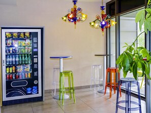 Máquina expendedora con aperitivos y bebidas, una mesita con taburetes y una planta en maceta cerca de una ventana.