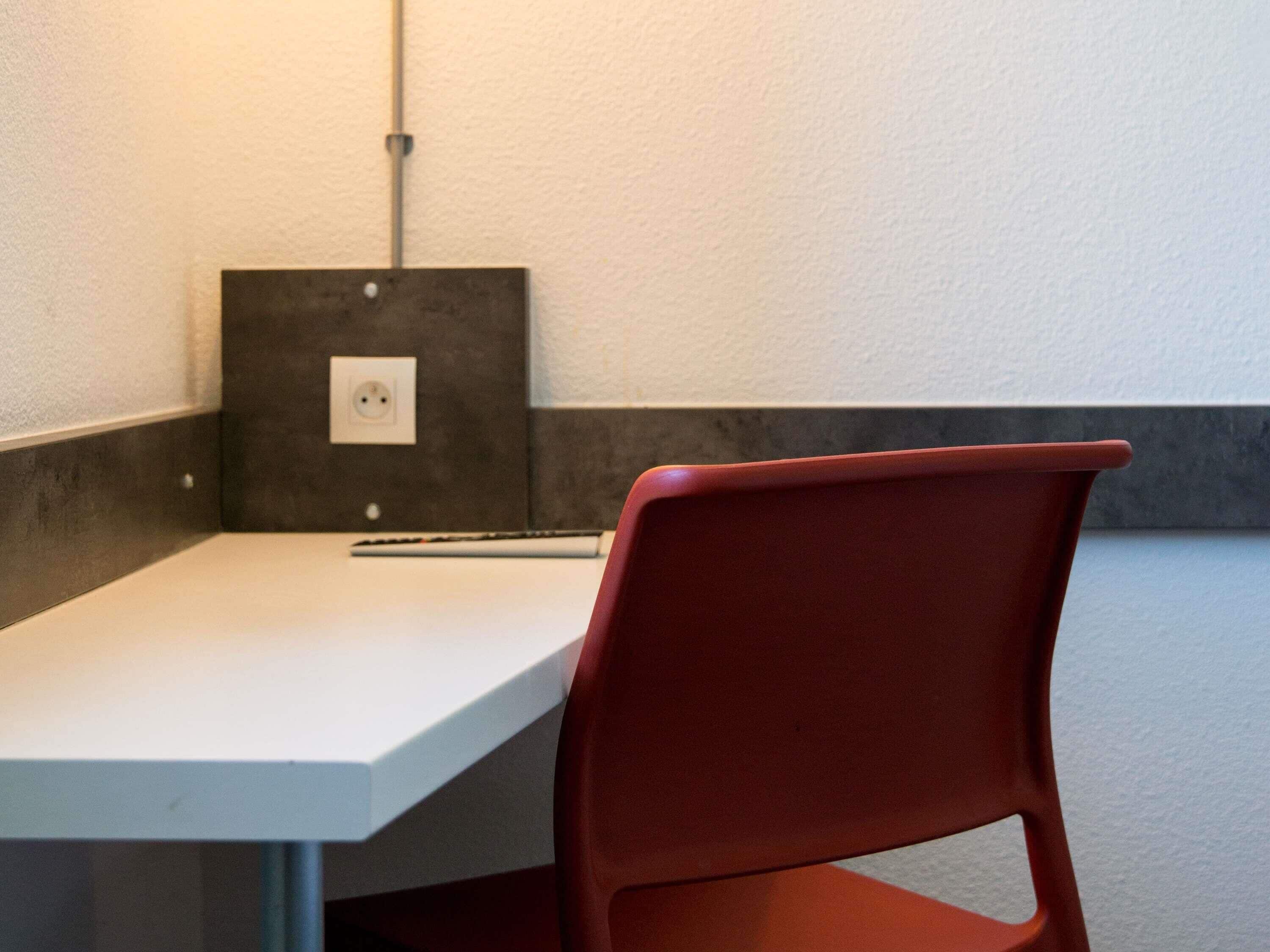 Une chaise rouge est placée devant une table blanche, près d’une prise de courant et d’un câble fixé au mur.