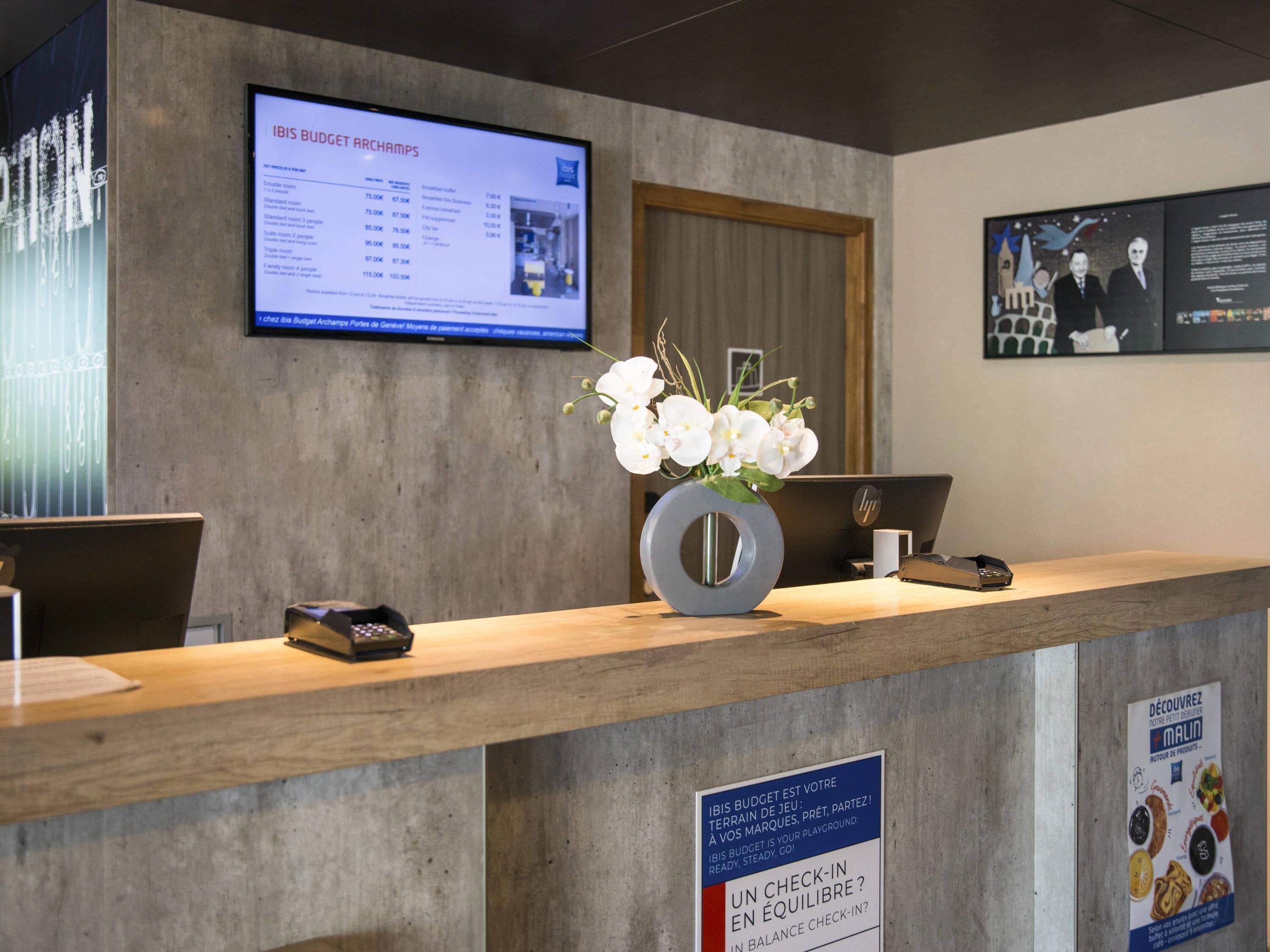 Un mostrador de recepción con un monitor que muestra un presupuesto, un florero con flores blancas y un letrero sobre el saldo de registro.
