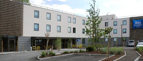 Un moderno edificio de hotel con tejado plano, múltiples ventanas y un letrero que dice 'Ibis Budget Hotels'. Hay zonas de estar al aire libre con sillas amarillas y pequeños árboles.