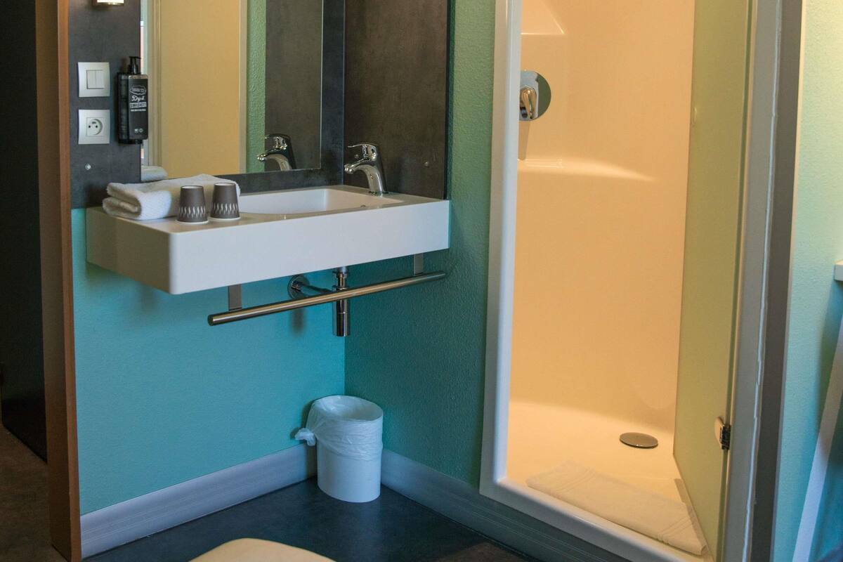 Un baño con lavamanos blanco, un espejo, toallero, cubo de basura y cabina de ducha.