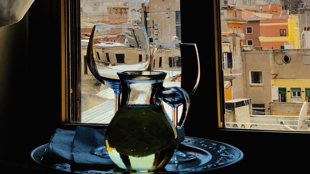 A view of a cityscape through a window with a table featuring a glass pitcher and a plate. - Art Atelier Vaticano (Rome)