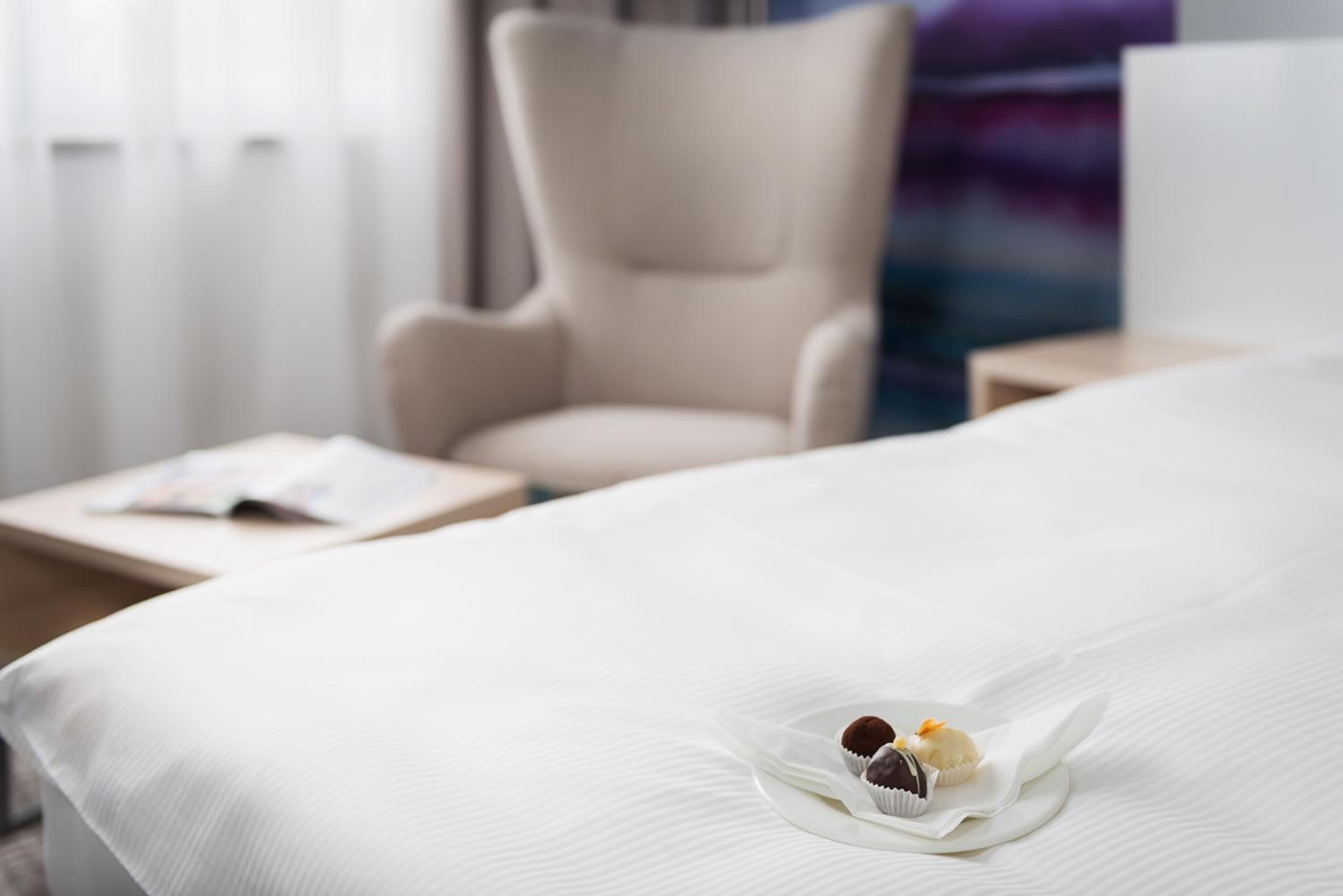 Una habitación de hotel con una cama bien hecha, una silla y una mesita con una revista.