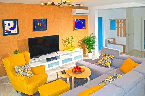 A modern living room with a flat-screen TV, yellow and grey sofas, and a coffee table with fruit.