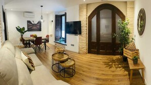 Una sala de estar moderna con un sofá, una mesa de comedor y sillas. Hay un televisor montado en la pared y una puerta de madera con un panel de vidrio.