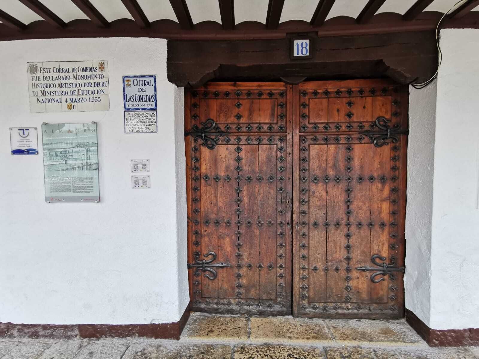 A wooden double door with metal hinges and decorative elements, set in a building with white walls and a dark wooden beam ceiling.