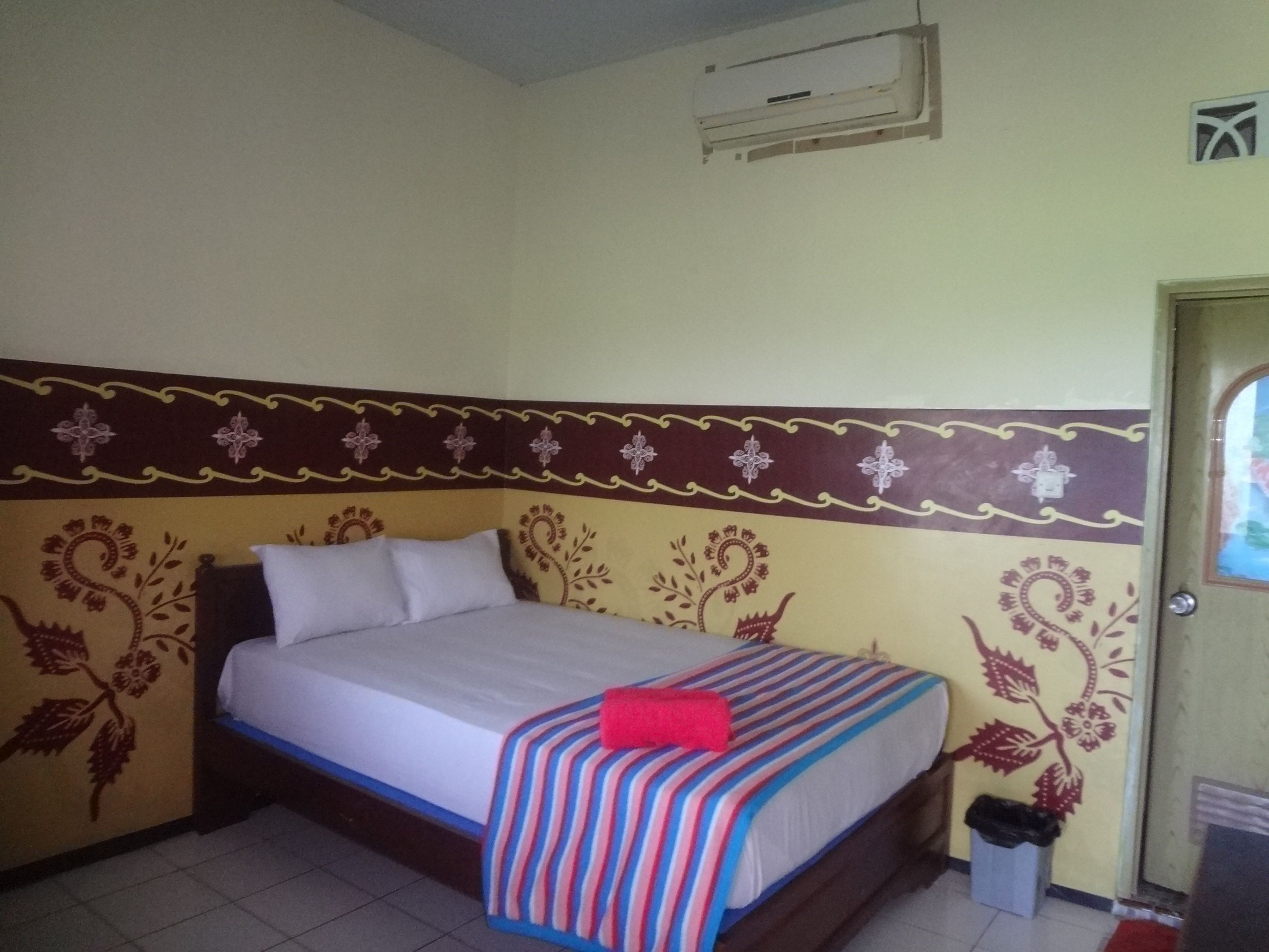 Una cama individual con una colcha y almohadas a rayas, un aire acondicionado en la pared y una puerta con ventana.