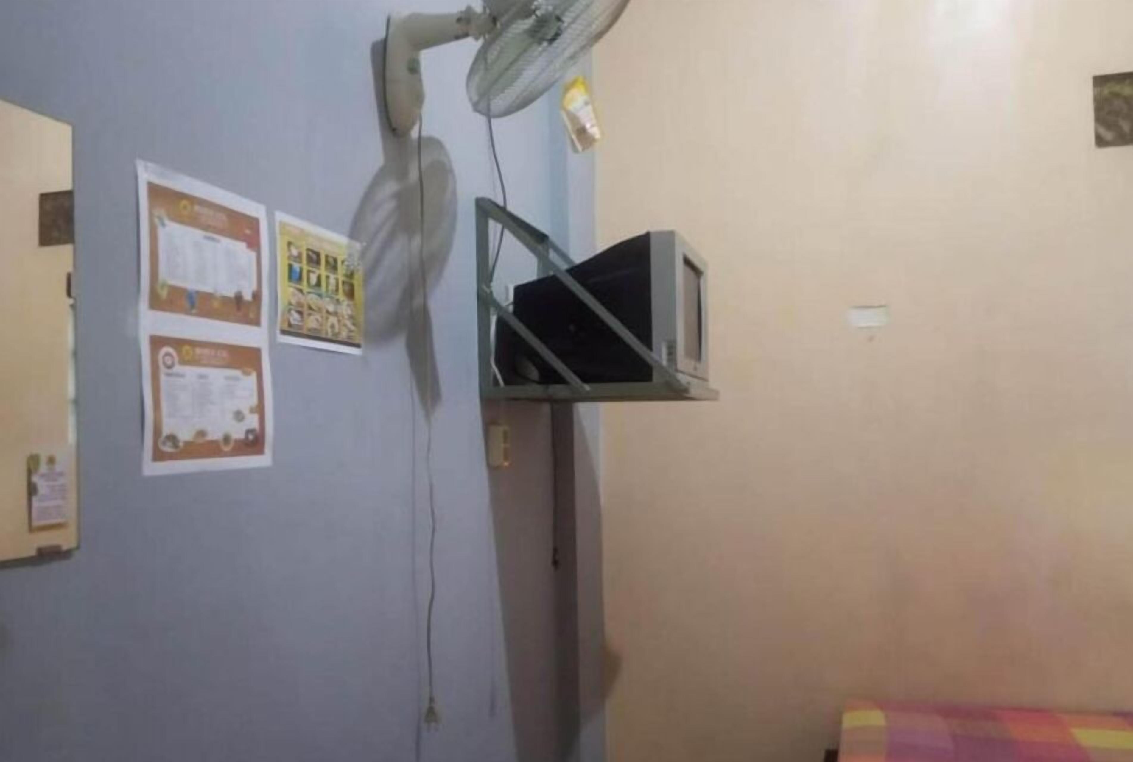 Una habitación con un ventilador de pared, un estante con un televisor y varios carteles en la pared.
