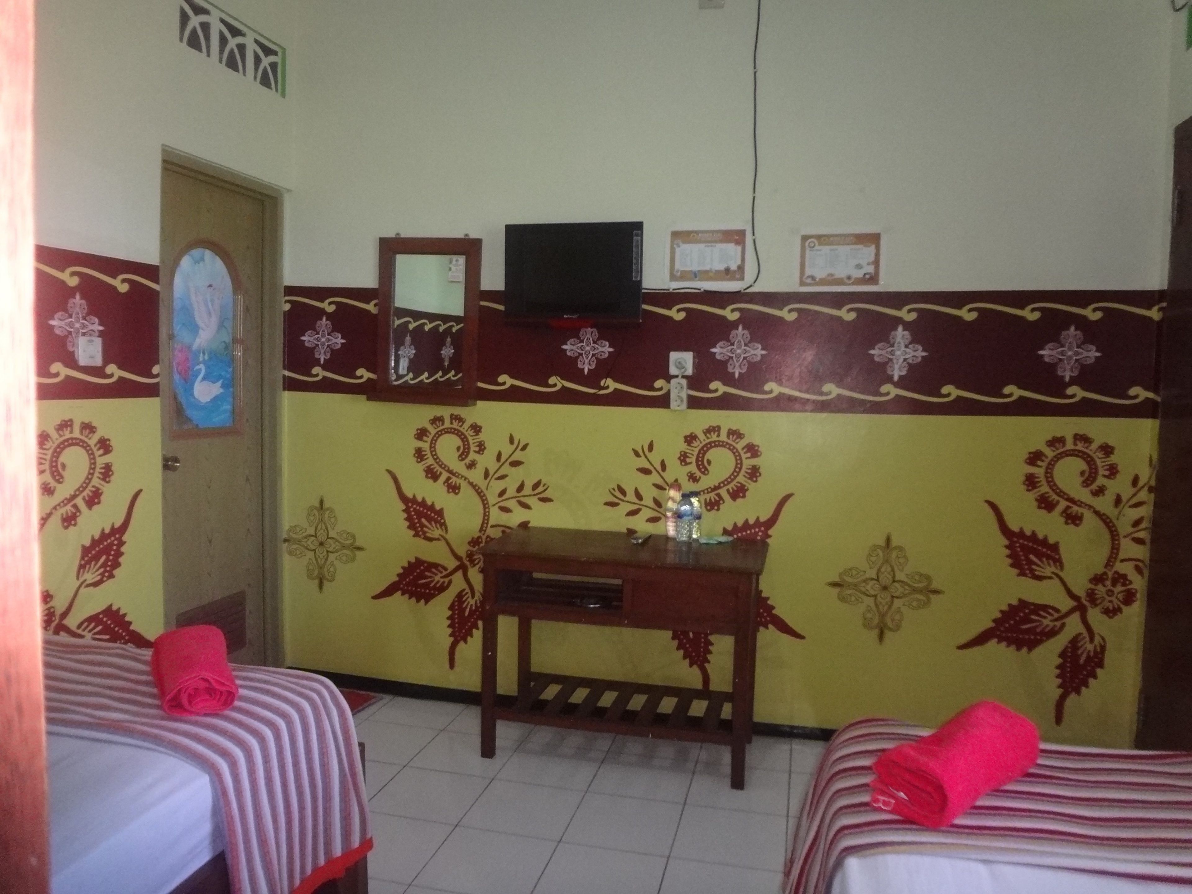 A room with two beds, a small table with a bottle, a TV on a stand, and decorative wall patterns.
