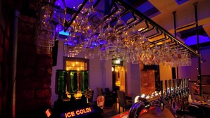 A bar area with a 'ICE COLD' dispenser, multiple glasses hanging from the ceiling, and a bar counter with a red mat. - George Wright Boutique Hotel, Bar & Restaurant. (Rotherham)
