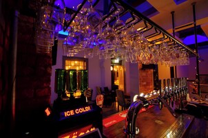 Uma área de bar com um dispensador 'ICE COLD', vários copos pendurados no teto e um balcão com um tapete vermelho.