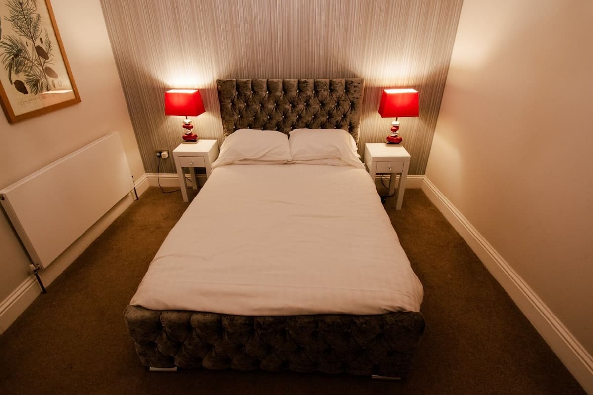 A neatly made bed with a tufted headboard, two bedside tables with red lamps, and a framed botanical print on the wall.