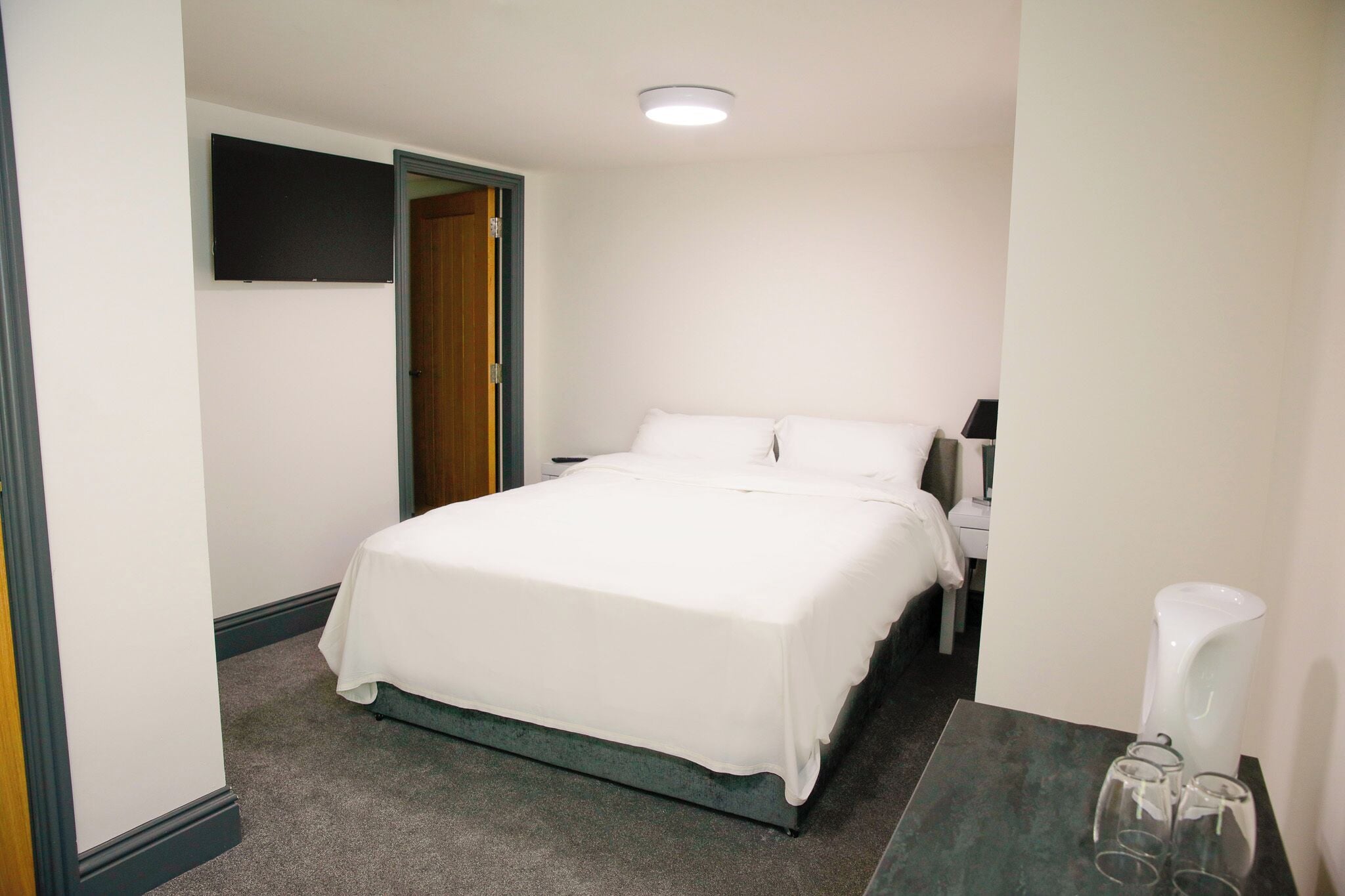 A bedroom with a bed, a nightstand, a lamp, and a bathroom area with a sink and mirror.