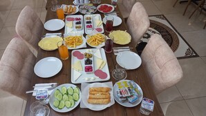 Mesa de comedor dispuesta con diversos alimentos y bebidas, rodeada de sillas con cojines.