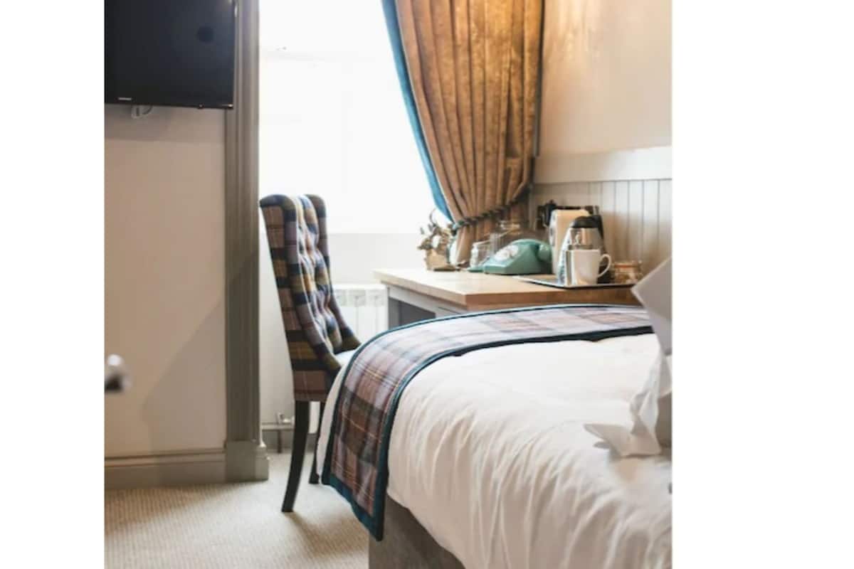 Quarto de hotel com cama, uma cadeira, escrivaninha com chaleira, cafeteira e televisão na parede.