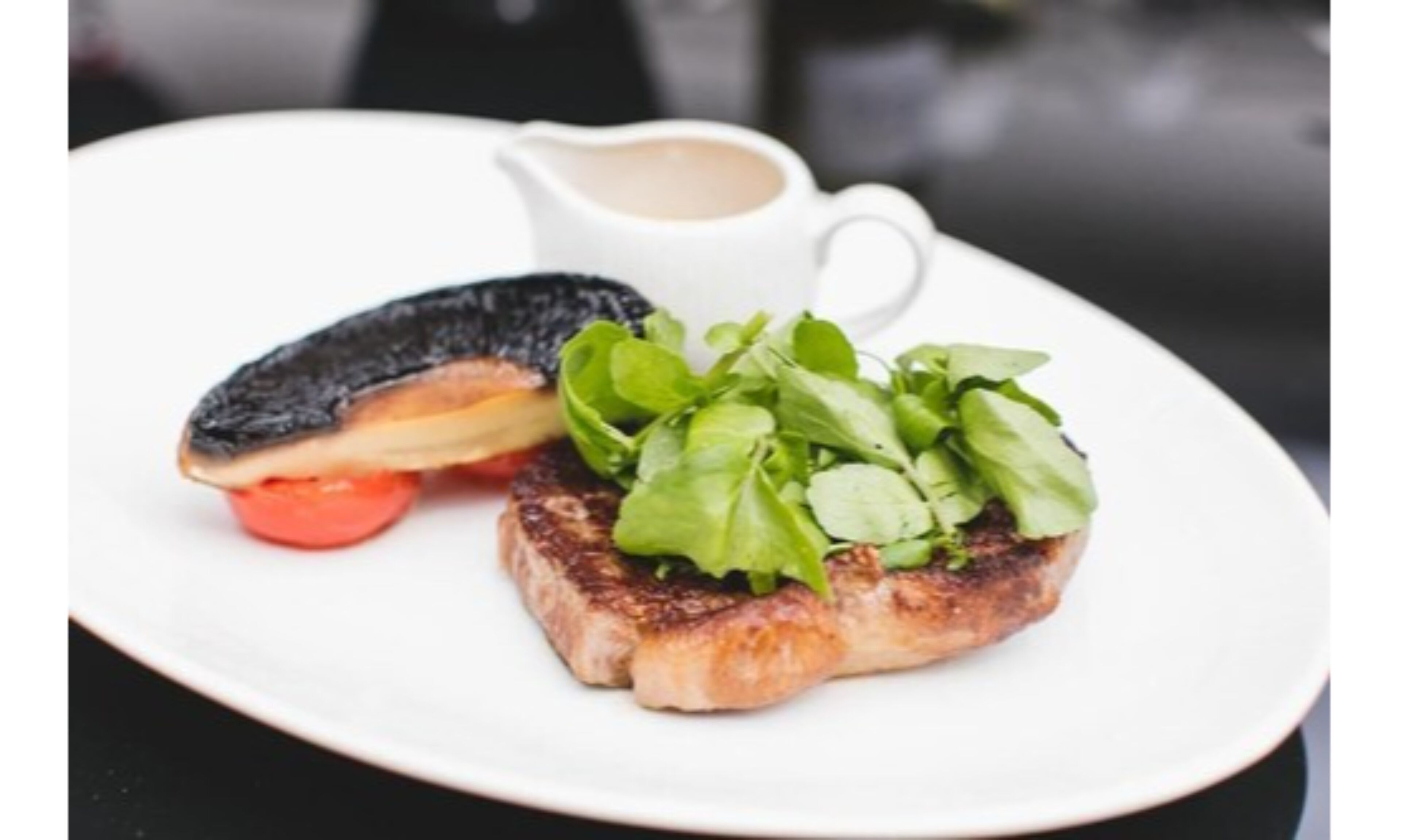 Un plato con un bistec a la parrilla, un tomate asado y una taza pequeña de salsa.