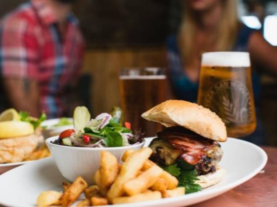 Un plato con una hamburguesa, papas fritas y una ensalada, y una jarra de cerveza al fondo.