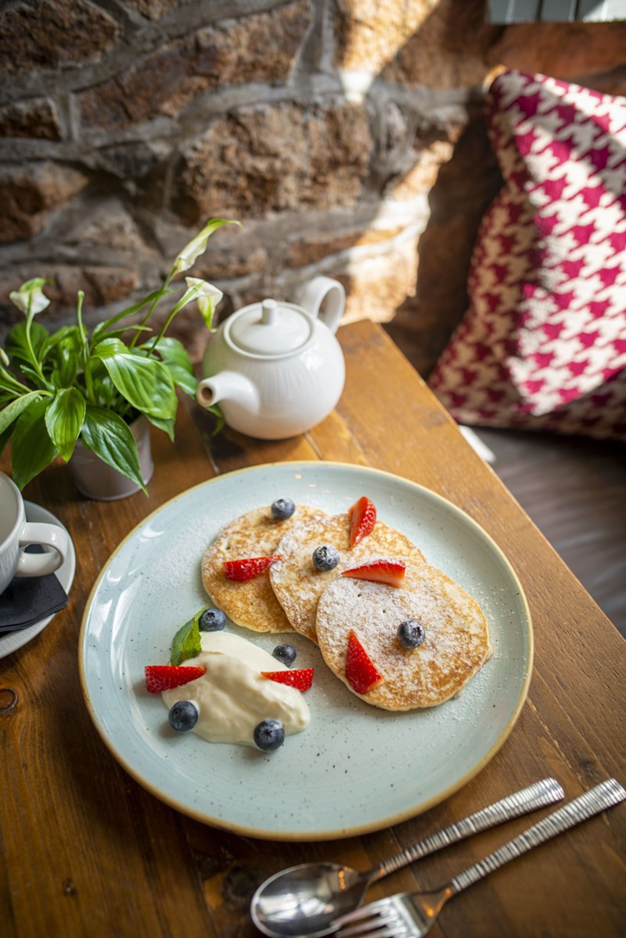 Een bord pannenkoeken met aardbeien en bosbessen, een theepot en een bloempot op een houten tafel.
