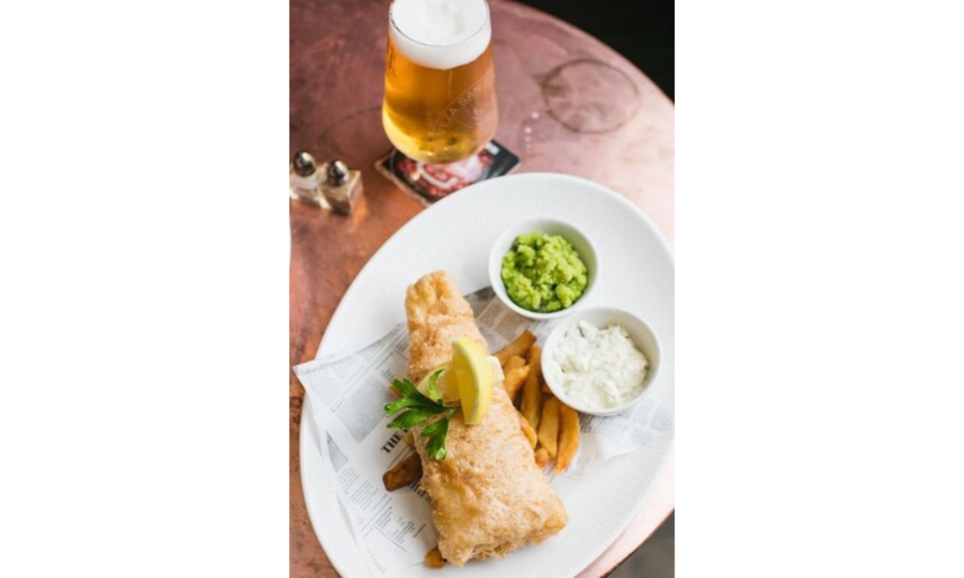 A plate with fish and chips, a glass of beer, and two side dishes.