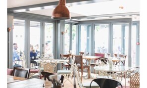 A restaurant interior with multiple tables and chairs, large windows, and hanging pendant lights.