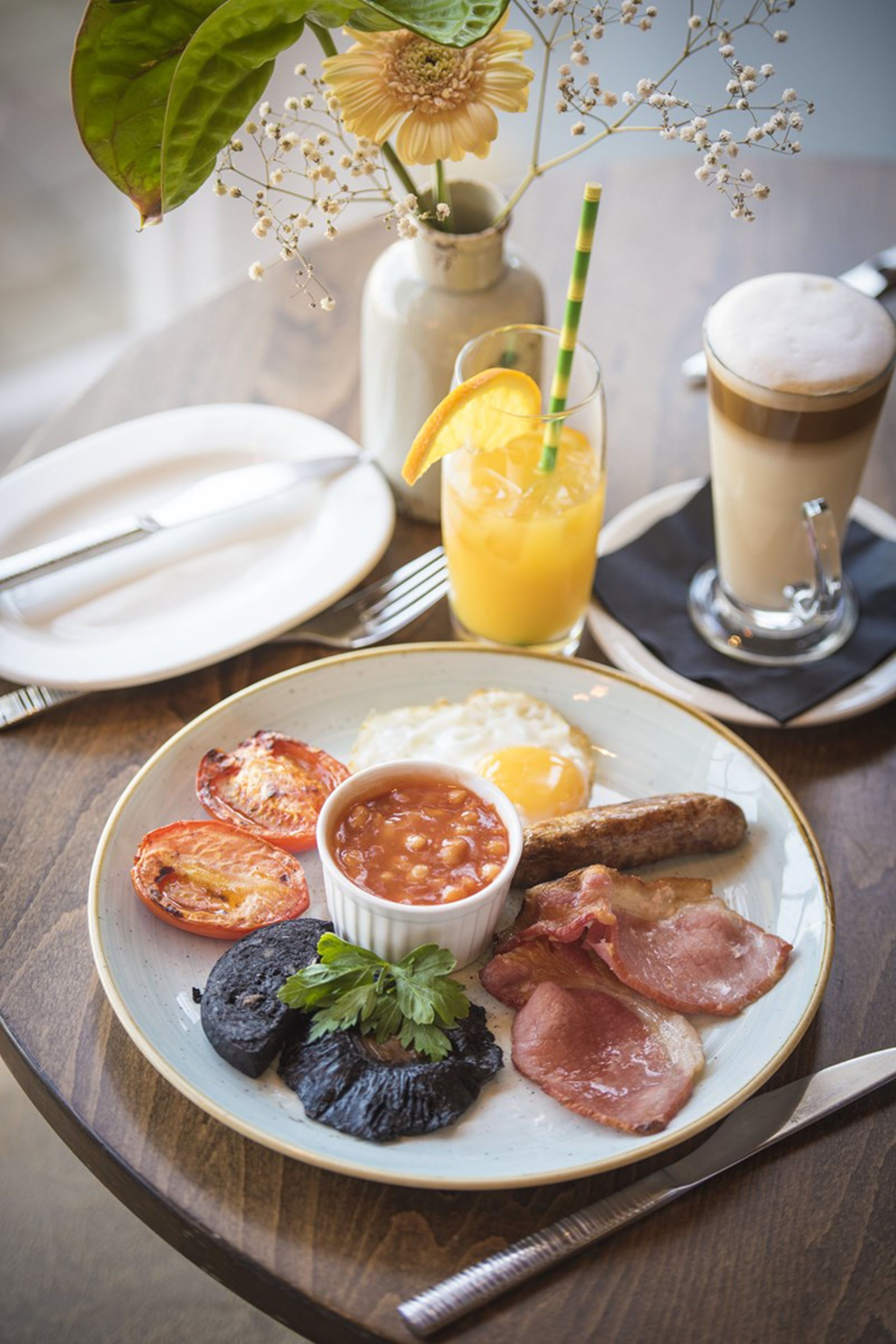 Un plato de desayuno con huevos, tocino, tomates y frijoles, un vaso de jugo de naranja y una taza de café sobre una mesa de madera.