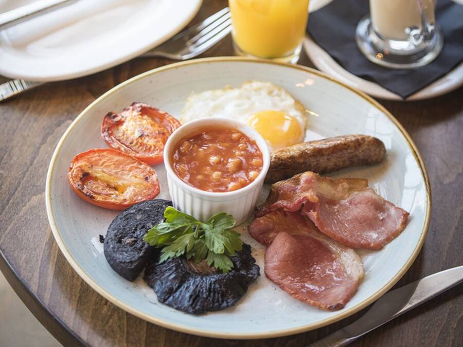 Un plato de desayuno con huevos, tocino, tomates y frijoles, un vaso de jugo de naranja y una taza de café sobre una mesa de madera.