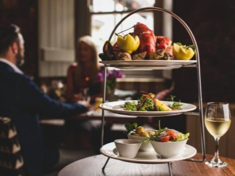 Un soporte de tres niveles con diversas frutas y pasteles, acompañado de una copa de vino blanco sobre una mesa.