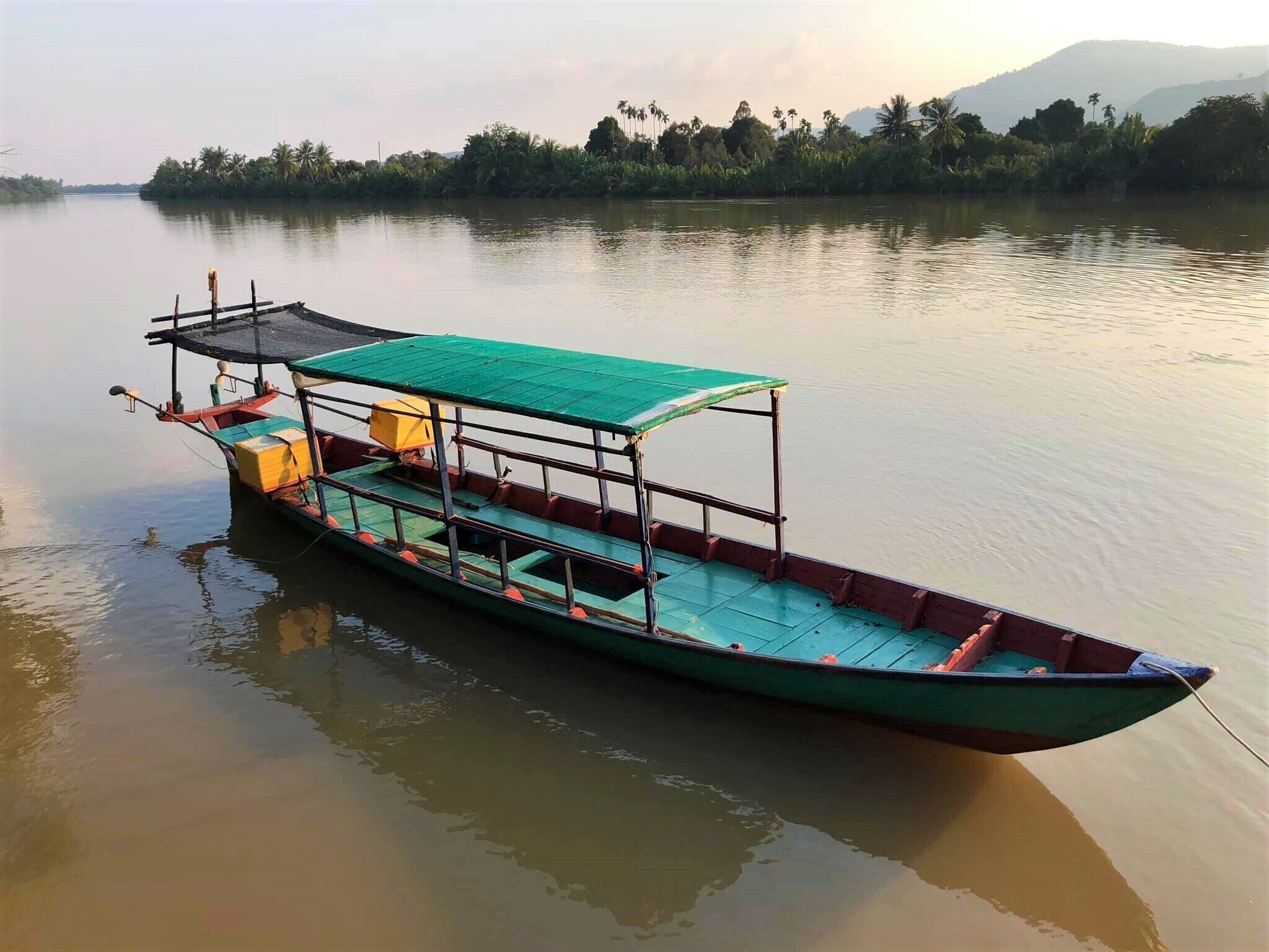 A long, narrow boat with a green canopy and multiple seating areas, docked on a calm river with a forested shoreline in the background.