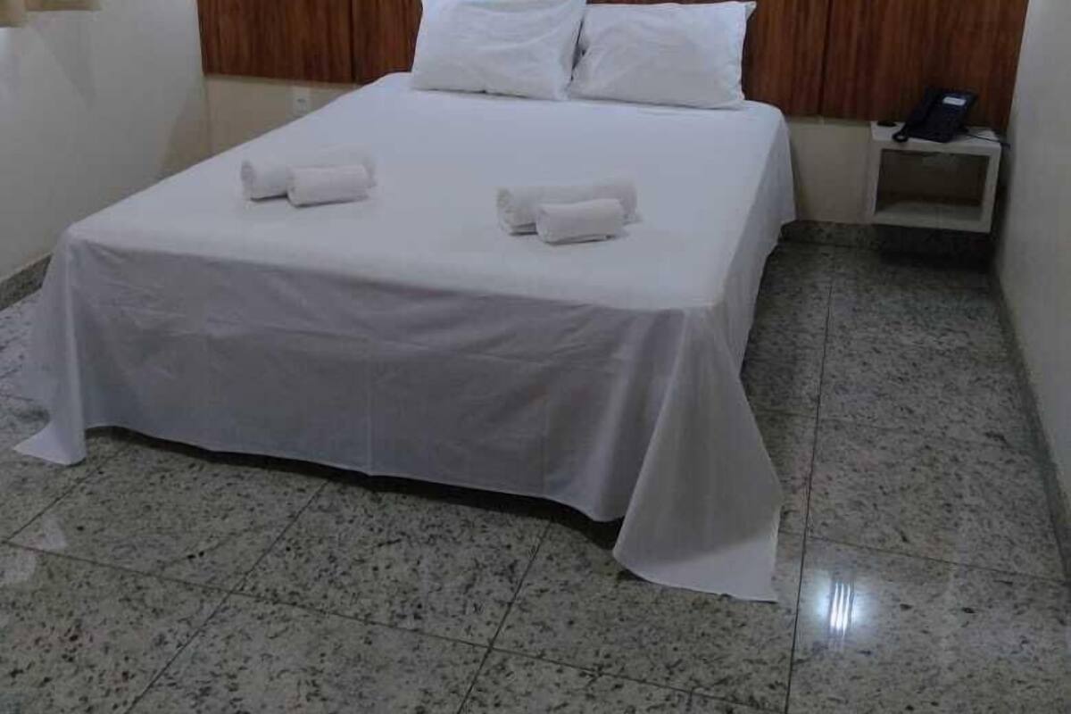 A hotel room with a bed, white linens, a wooden headboard, two bedside lamps, a nightstand with a phone, and a tiled floor.
