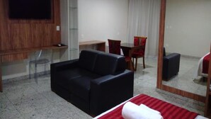 A hotel room with a black sofa, a small desk, a chair, a television mounted on a wooden panel, and a large mirror. - Topazio Imperial Hotel (Itauna)