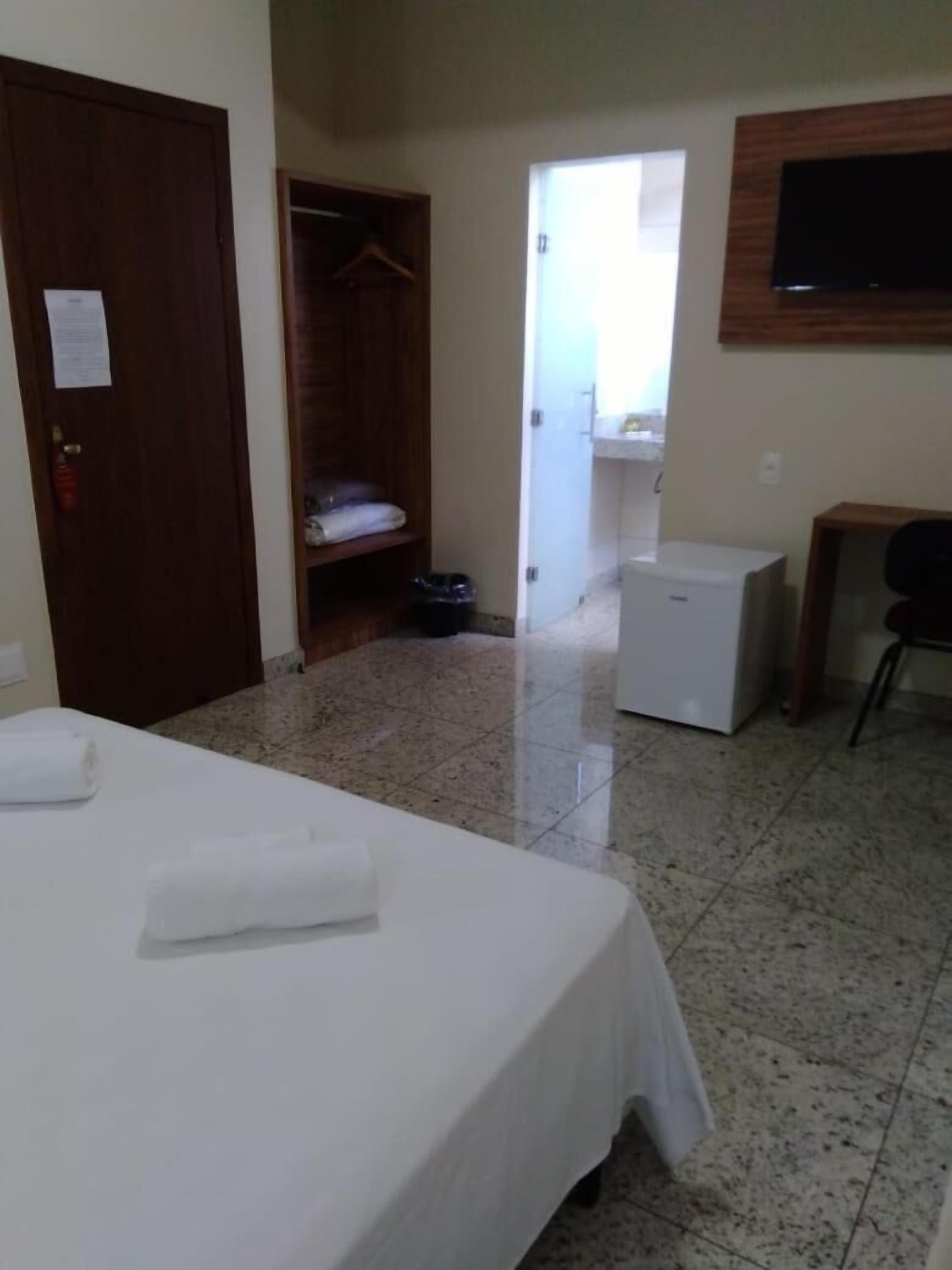 A hotel room with a bed, a small refrigerator, a TV mounted on the wall, and a closet with hanging clothes.