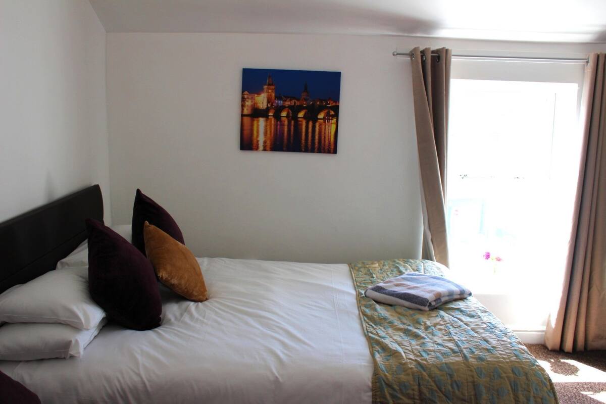 A bedroom with a bed, pillows, a blanket, a painting, and a window with curtains.